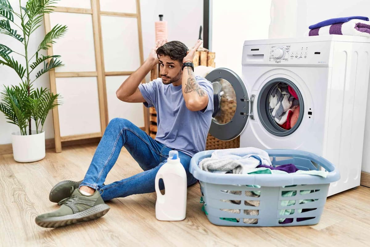 A young man is seated beside a load of laundry as he tries to figure out the safest screen-printing washing technique to properly clean and protect the prints on his favourite custom tees.